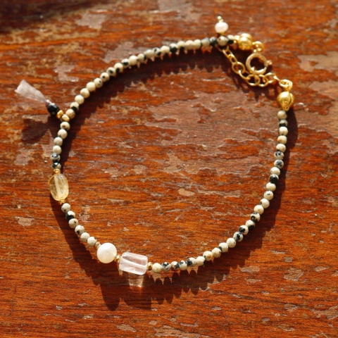 Bransoletka z drobnymi koralikami w ozdobiona kilkoma większymi kamieniami o różnych barwach na drewnianym tle.