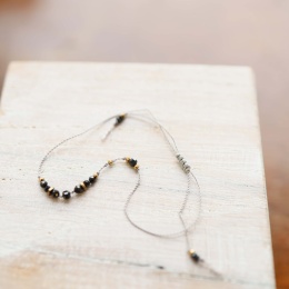 Bransoletka na szczęście z czarnym turmalinem na szarej nylonowej nici z delikatnym złotym wykończeniem, ułożona na jasnym tle.