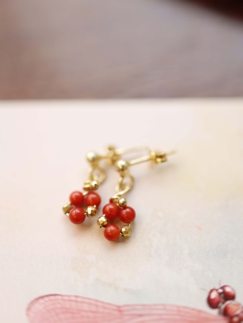 Złote kolczyki z czerwonym koralem na tle ilustracji w pastelowych kolorach, zdjęcie biżuterii