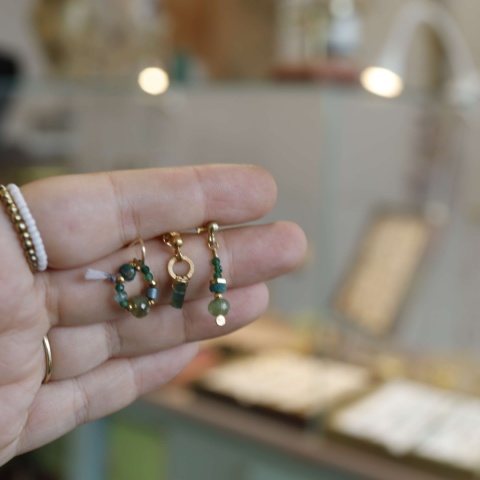 Zestaw trzech różnych kolczyków z serii „Pary nie do pary”, z elementami z zielonych kamieni i złotymi detalami na tel dłoni
