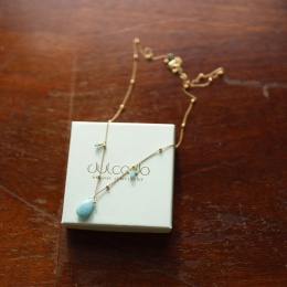 Złocony naszyjnik z kuleczkami oraz zawieszkami z amazonitu i opalu, zdjęcie na tle opakowania z logo marki.